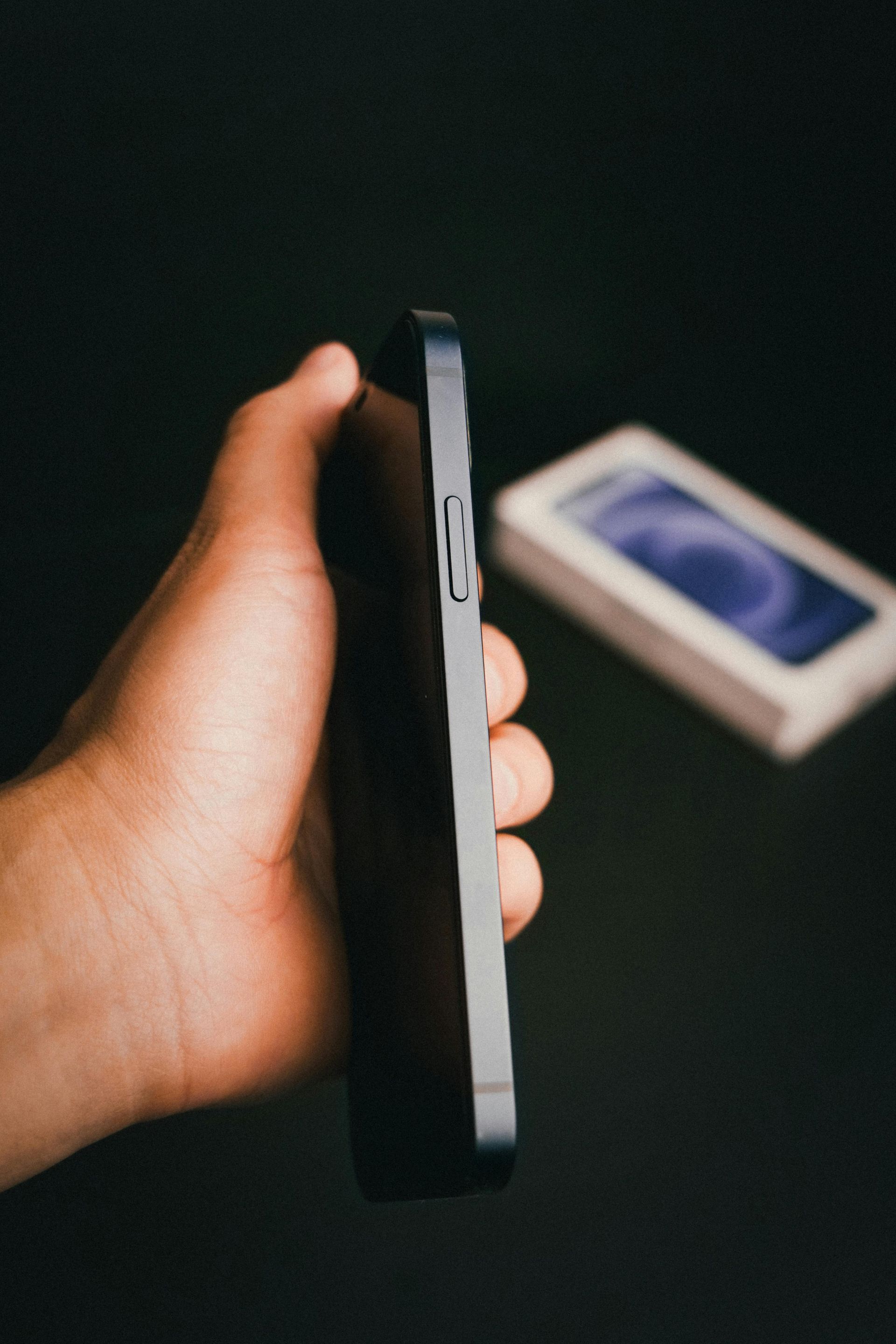 Eine Hand hält ein dunkelblaues Smartphone, Seitenansicht, im Hintergrund die Verpackung des Telefons, das auf einer dunklen Oberfläche steht.