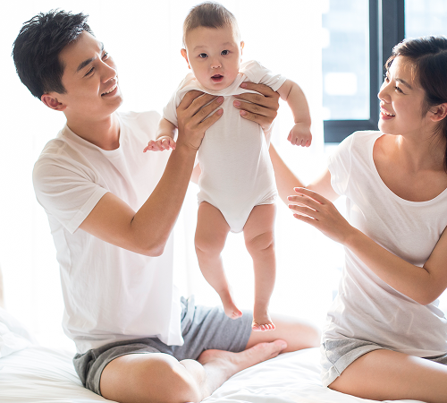 Asian parents lifting baby in the air, smiling. They are indoors, on a bed.