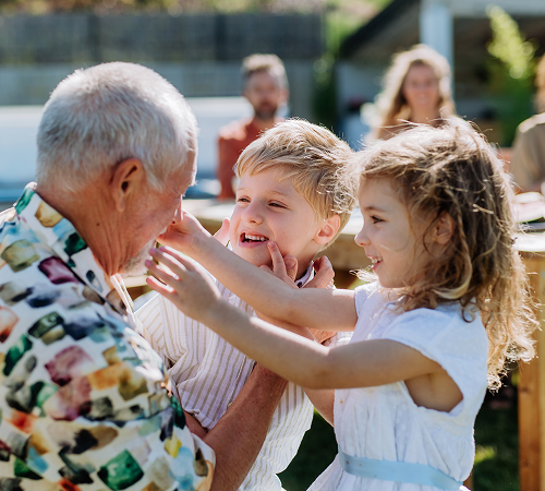 A grandfather is embraced by two young children outdoors; smiles and joy are evident.