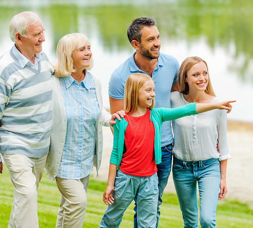 Family of six smiling and walking outdoors by a lake. A girl points right.