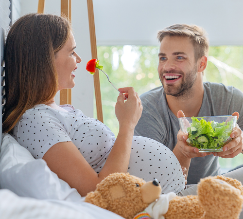 Pregnant woman in bed, smelling a flower, man offering salad. Happy couple in a bright room.