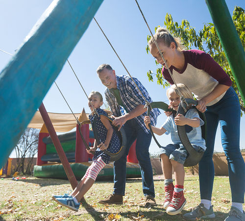 Family pushing two children on swings in a park, sunny day.