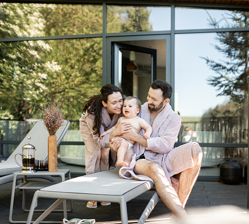 Family in bathrobes on a patio, smiling at a baby. Green trees in background.