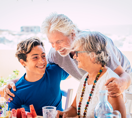 Grandparents and grandson smiling, outdoors. Older man's arm around the teen, woman wearing necklace.