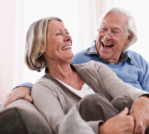 Smiling senior couple on a couch. The woman is wearing a cardigan, the man glasses.