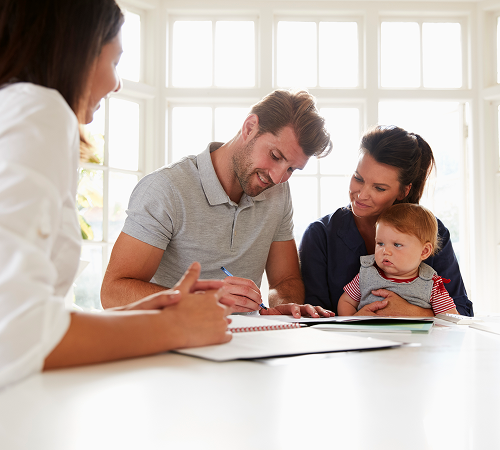 A woman, couple, and baby at a table, signing documents in a bright room.
