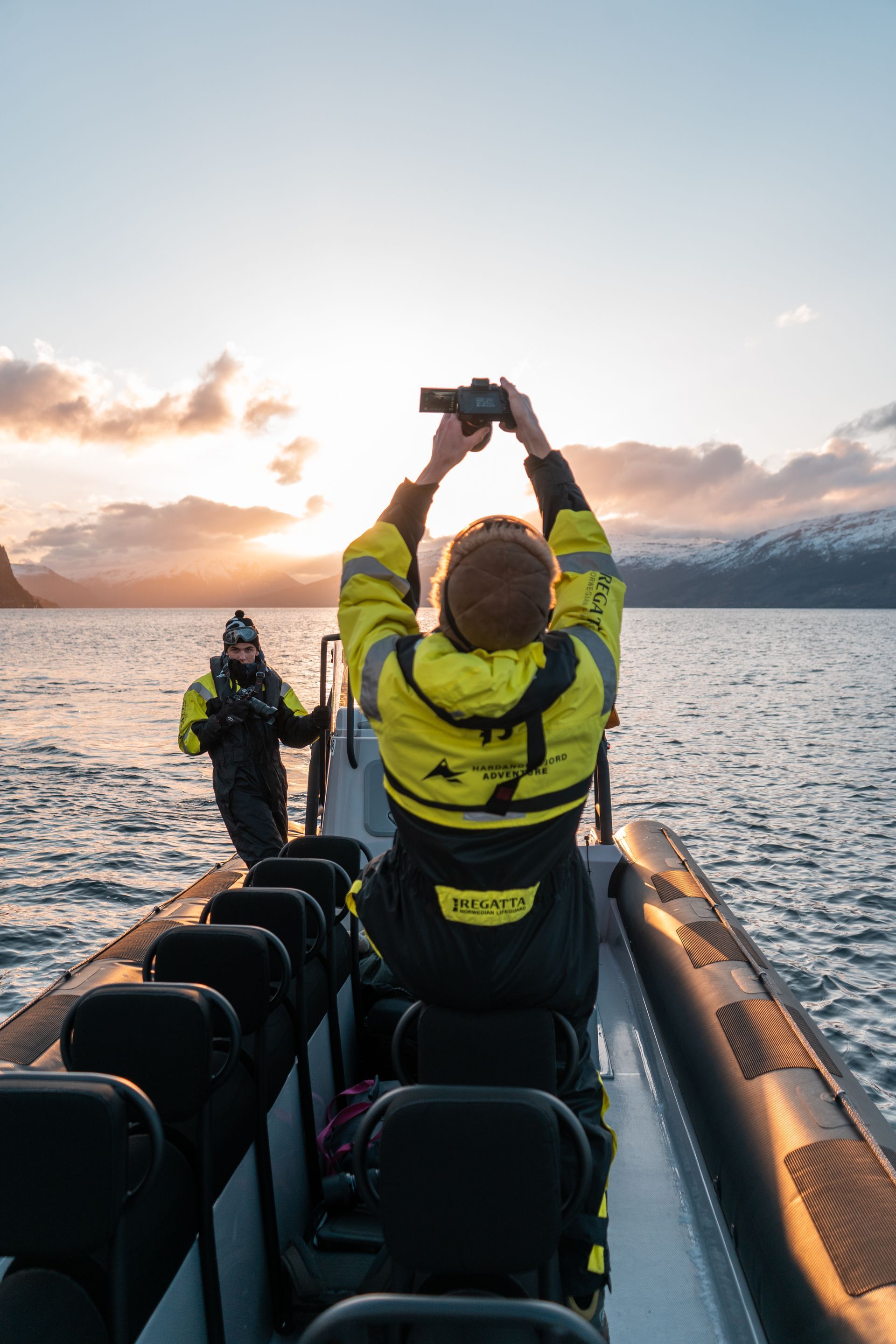 Two people in yellow suits on a boat, taking photos with a sunset backdrop.