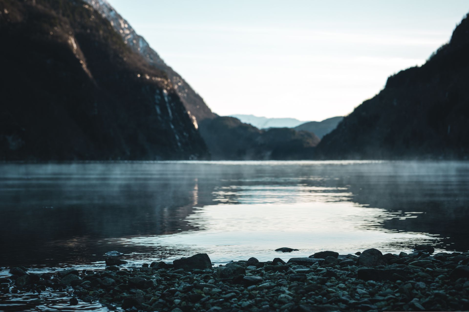 Calm lake between dark mountains; fog hovers above water; sunny sky.