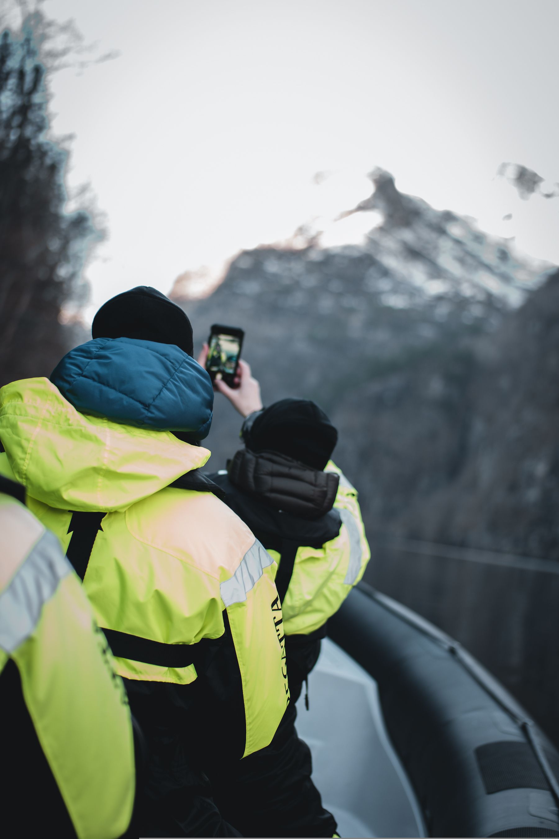 People in yellow jackets taking a photo of a snow-capped mountain from a boat.