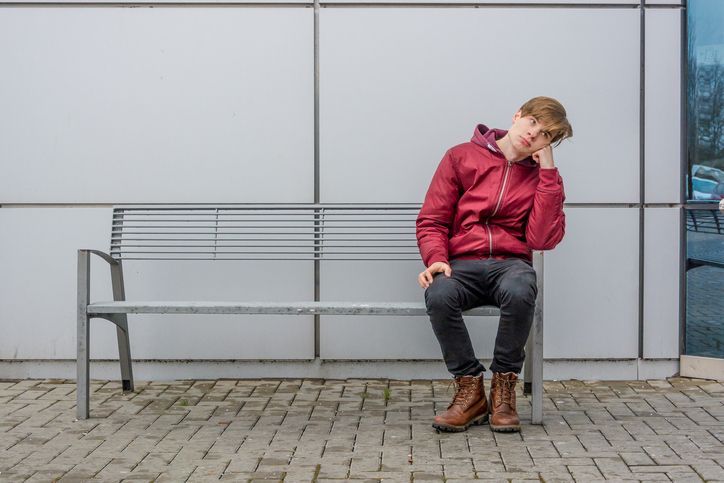 A teenage boy waits on a bench looking bored.
