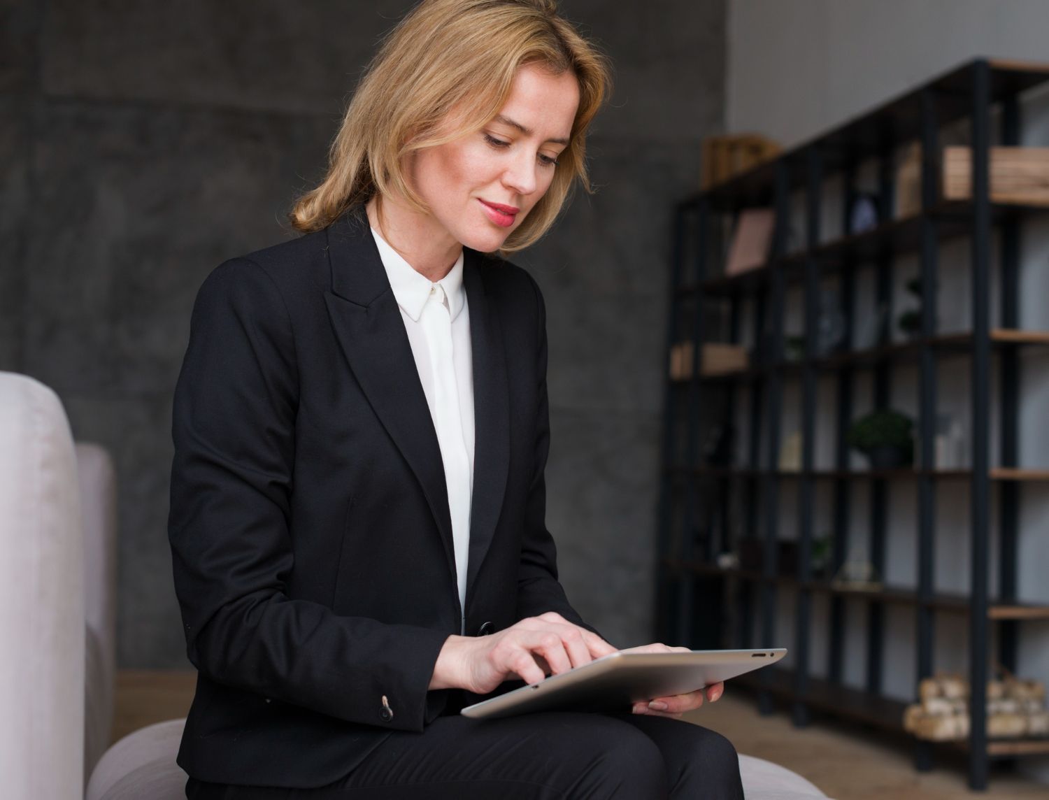Woman in a black suit uses a tablet, seated indoors with a bookshelf in the background.