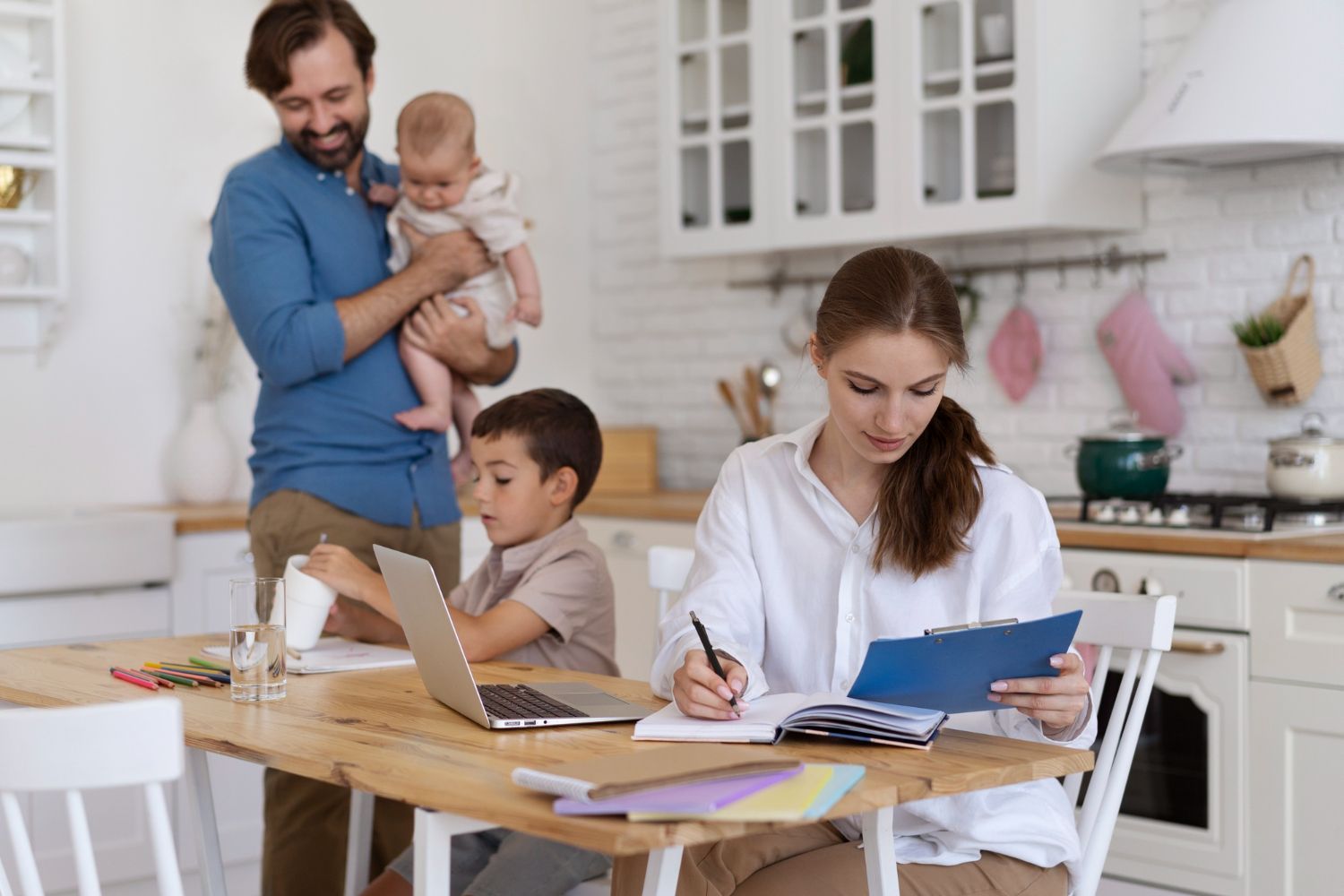 Family in a kitchen. Mother working on paperwork, father holding baby, boy at laptop. Bright setting.