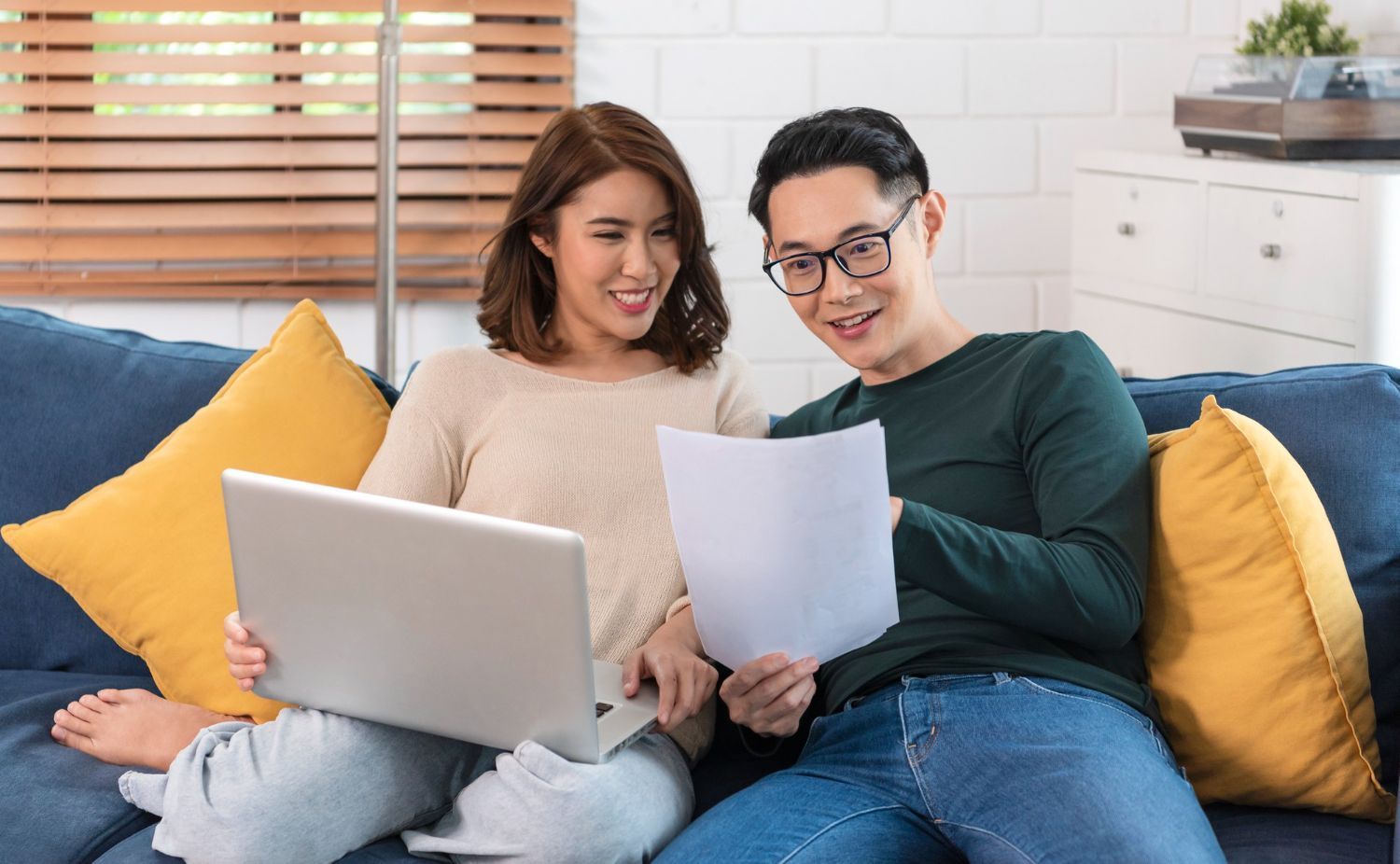 Couple on a blue couch, looking at a laptop and documents; smiling. Room with natural light, yellow pillows.