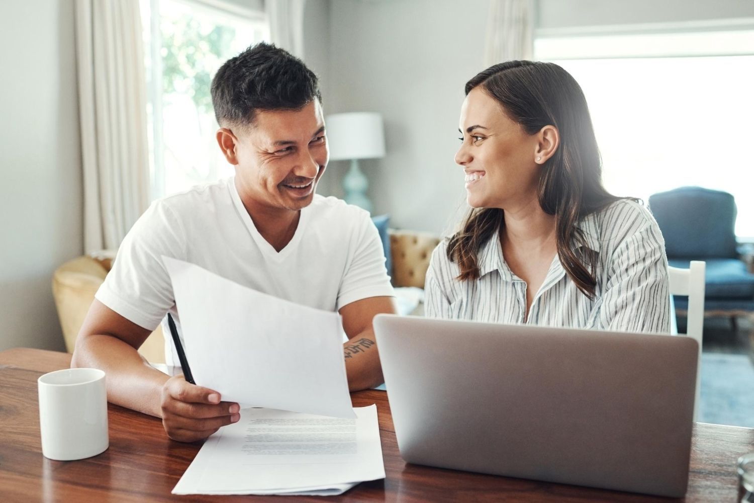 Couple smiling, reviewing documents and using a laptop at a table in a home.