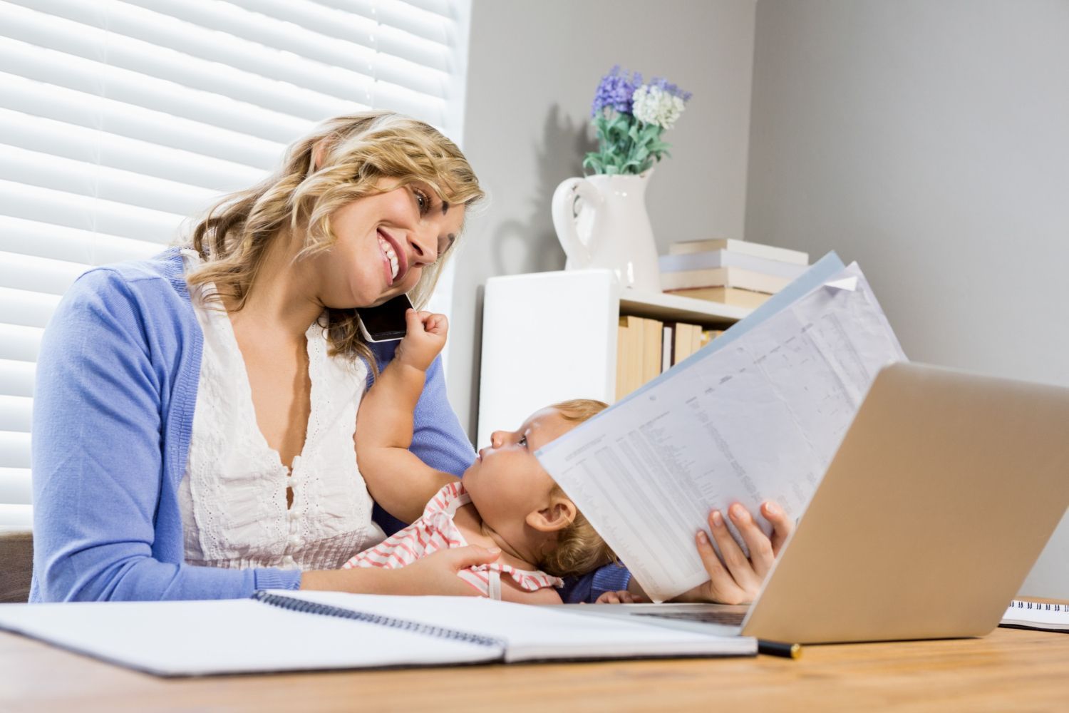 Woman on phone smiles, holding a baby. Working at a desk with laptop and paperwork.