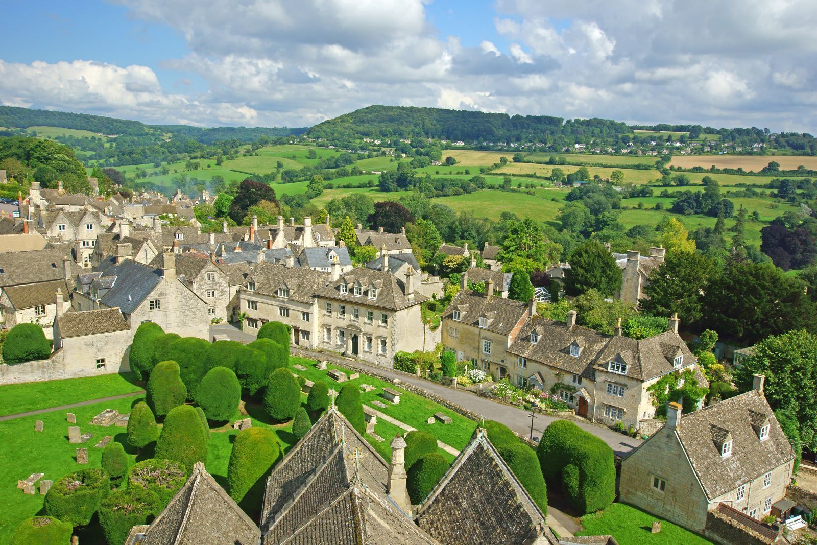 A scenic aerial view of Cotswold village with stone buildings, green fields, and a cloudy sky.