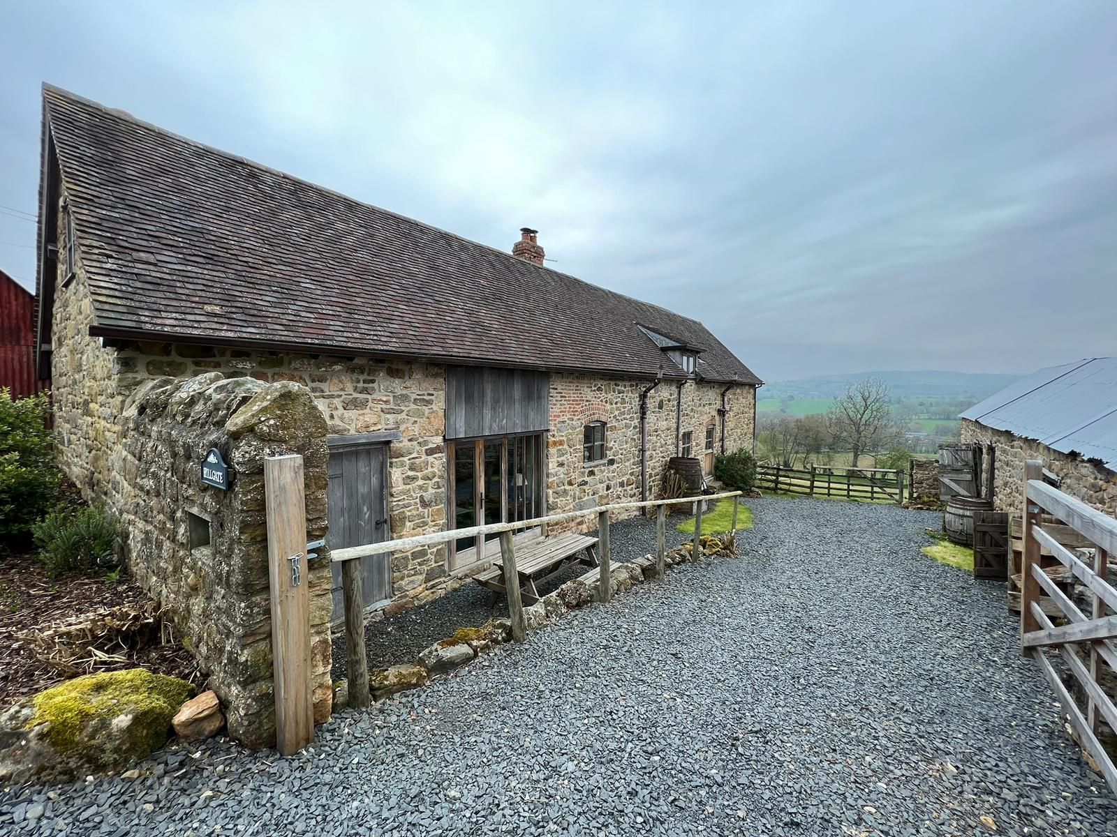 Stone barn with gravel driveway on a cloudy day, overlooking a green landscape.