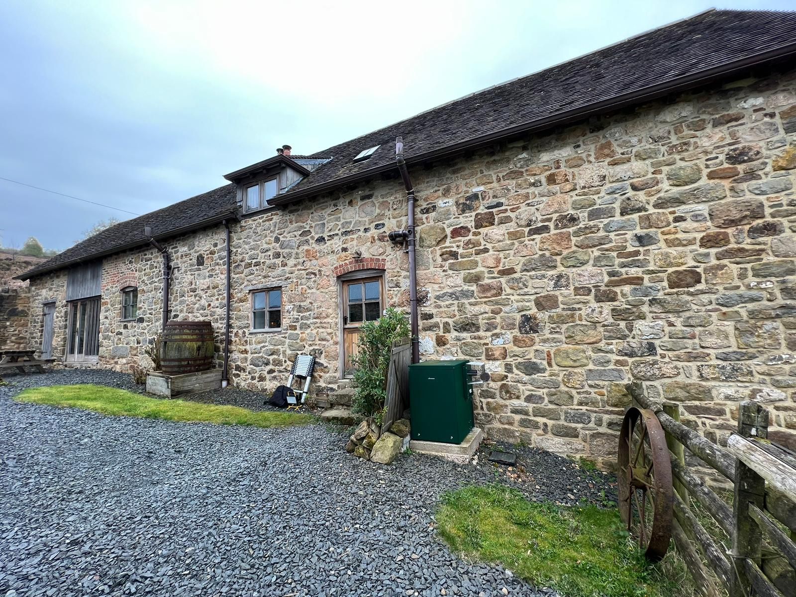 Stone barn under cloudy sky; gravel path, water tank, and wooden fence in foreground.