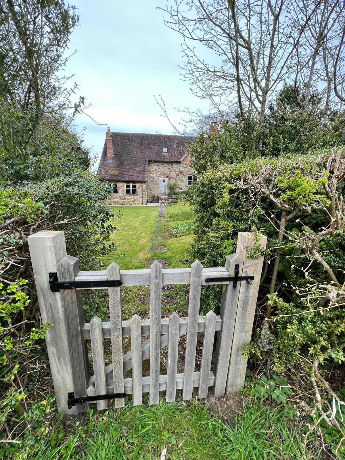 Wooden gate leading to a stone house, framed by green hedges and trees under a cloudy sky.