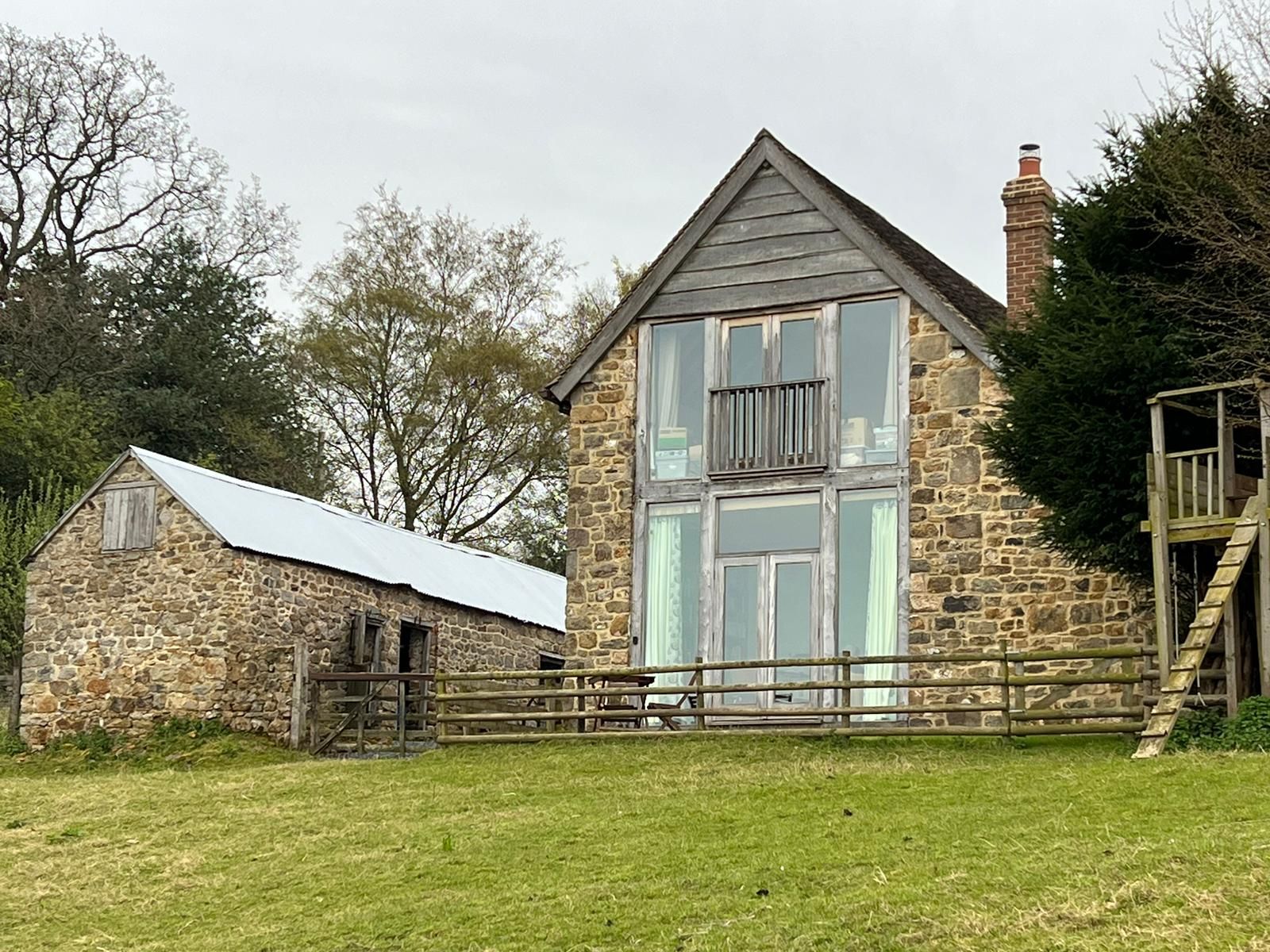 Stone buildings with a large window and a smaller barn, set on a grassy hill with a cloudy sky.