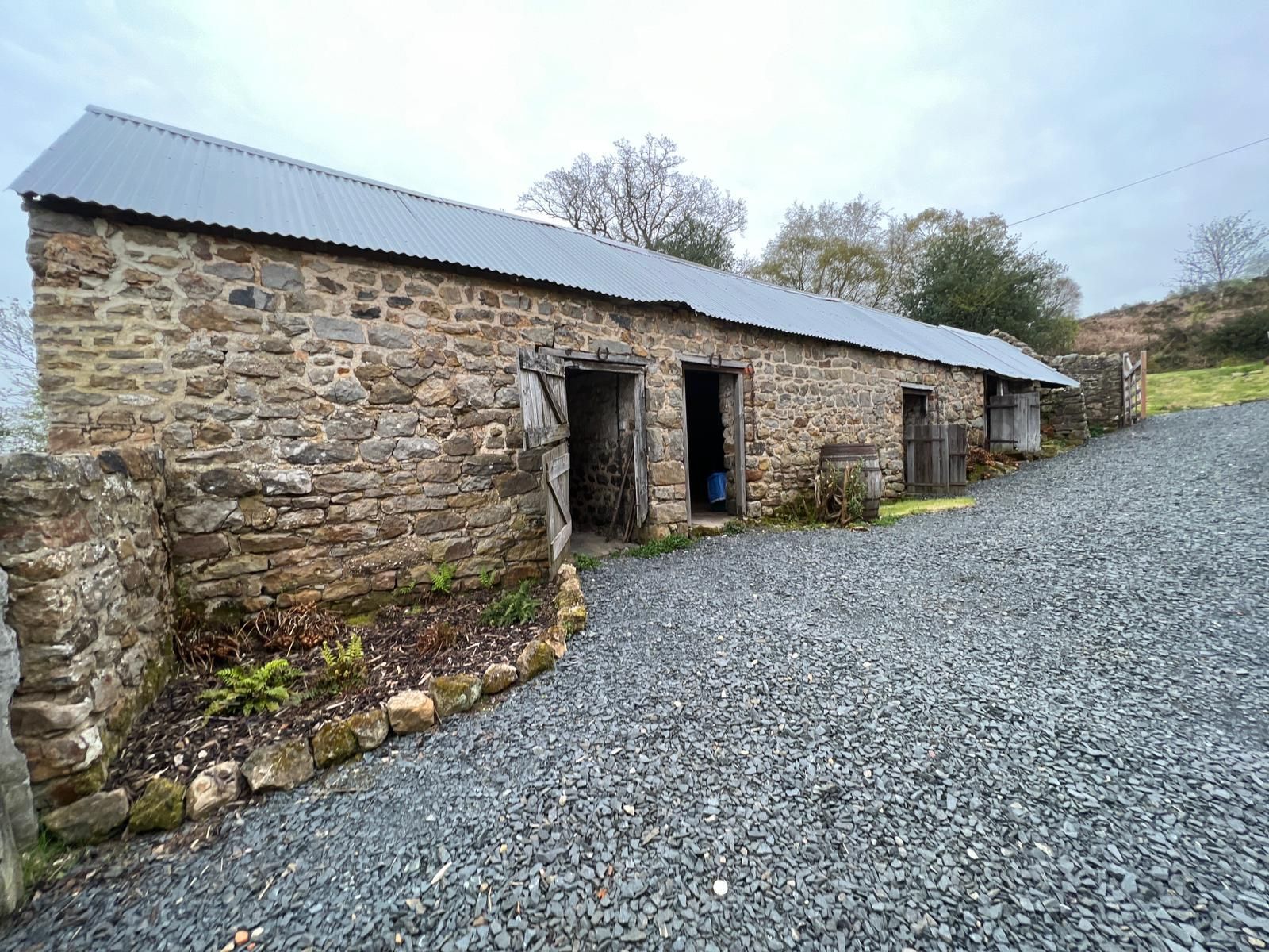 Stone building with open doorways, gravel driveway, under cloudy sky.
