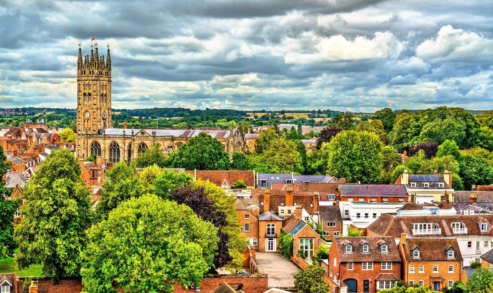 View of Tewkesbury Abbey tower and red-roofed buildings, trees, and cloudy sky.