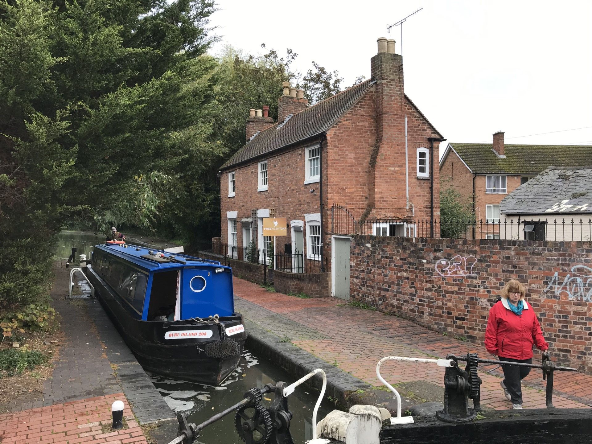 Canal boat in lock, red brick building, person in red jacket, trees, cloudy sky.