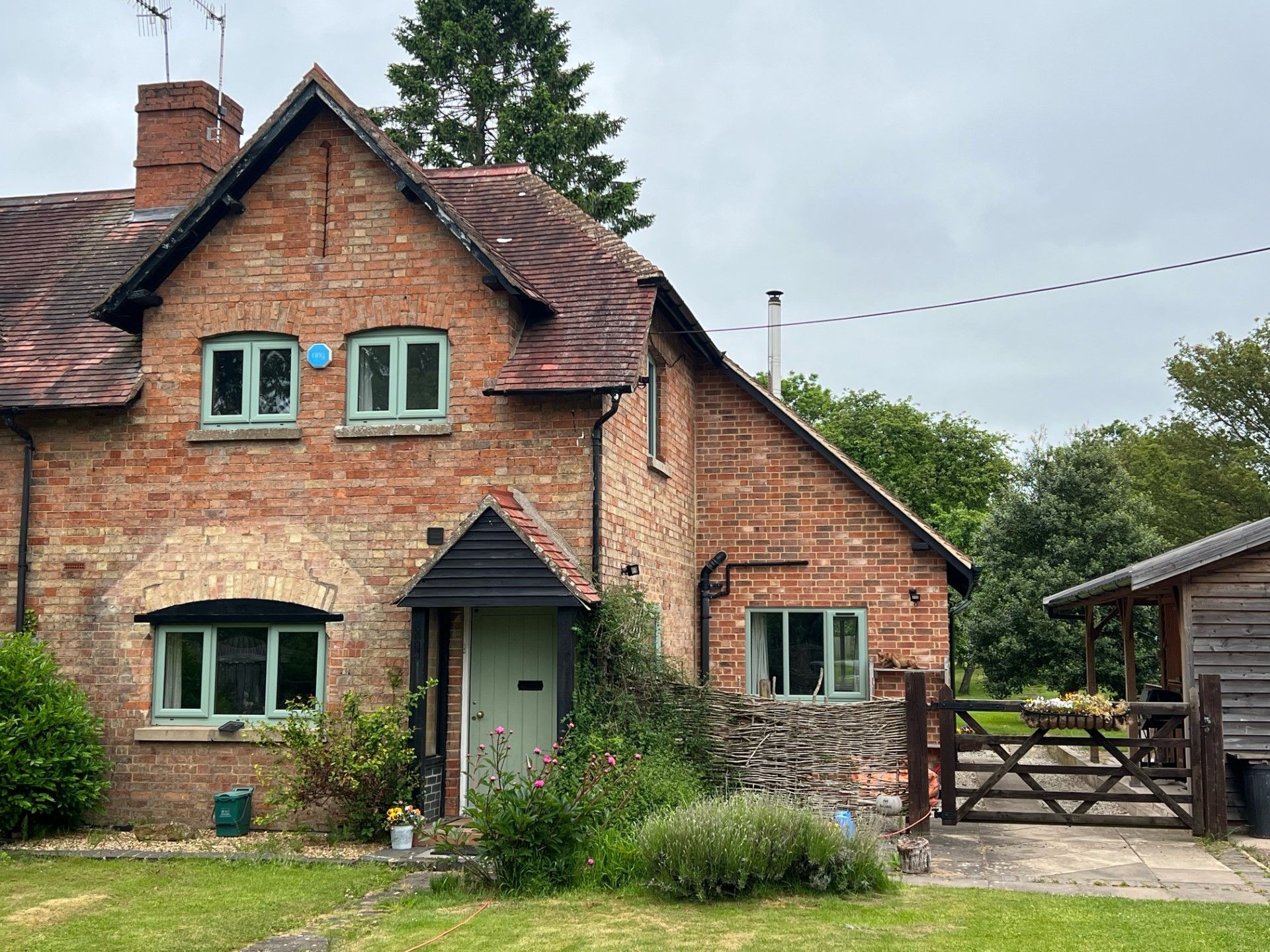 Red brick cottage with green door and windows, small wooden gate, and cloudy sky.