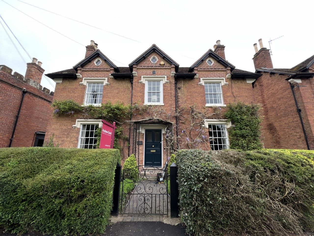 Red brick house with ivy, windows, and a dark door. Green hedge in front.