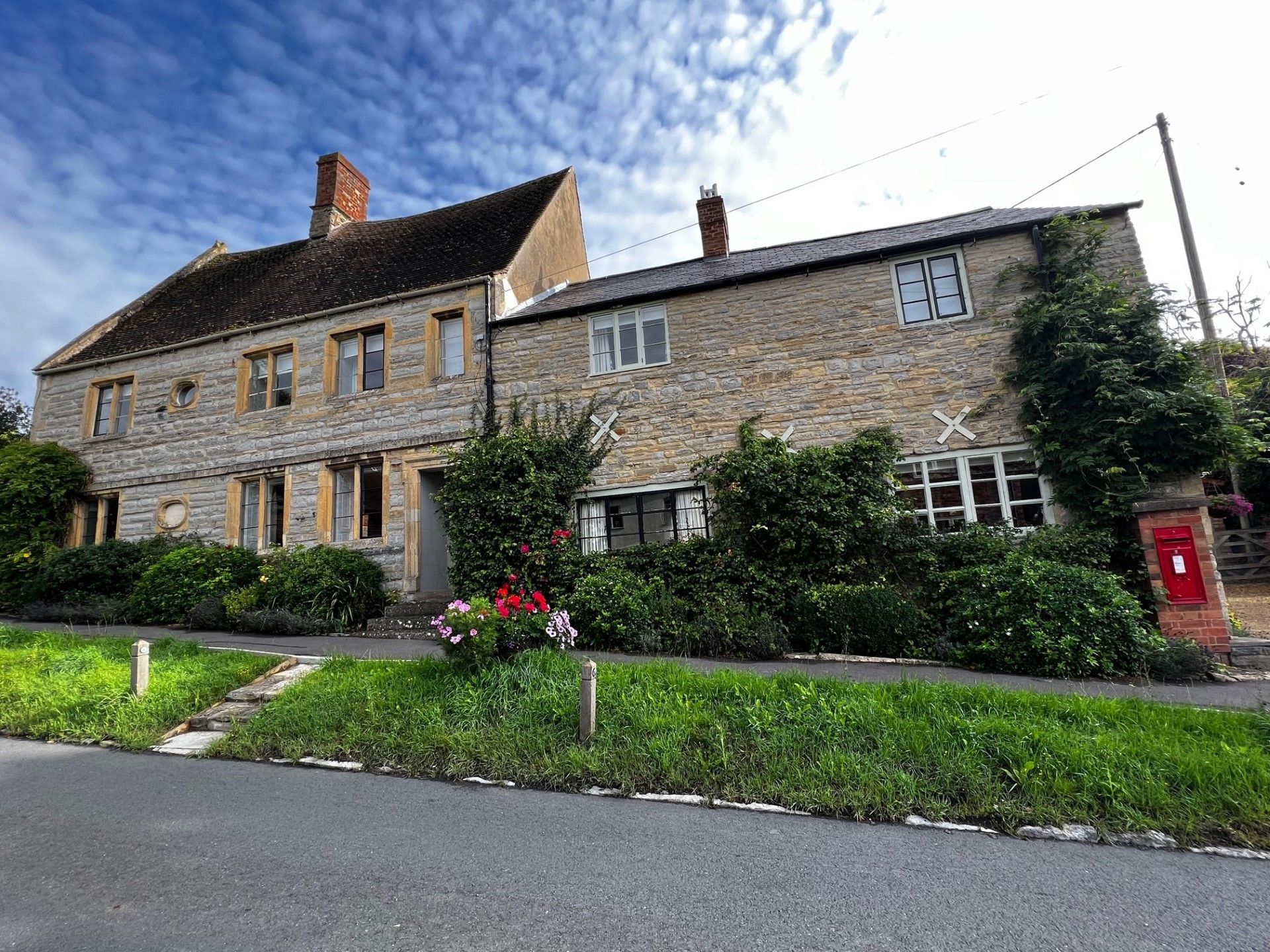 Stone buildings with windows, a red postbox, and flowering bushes line a road, under a blue sky.