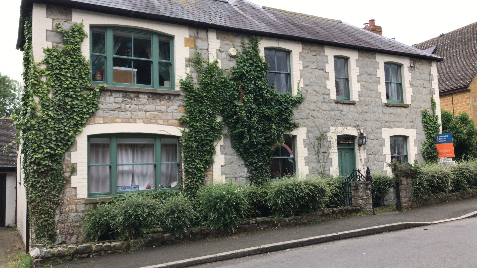 Stone house with green windows and door, partially covered in ivy. For sale sign visible.