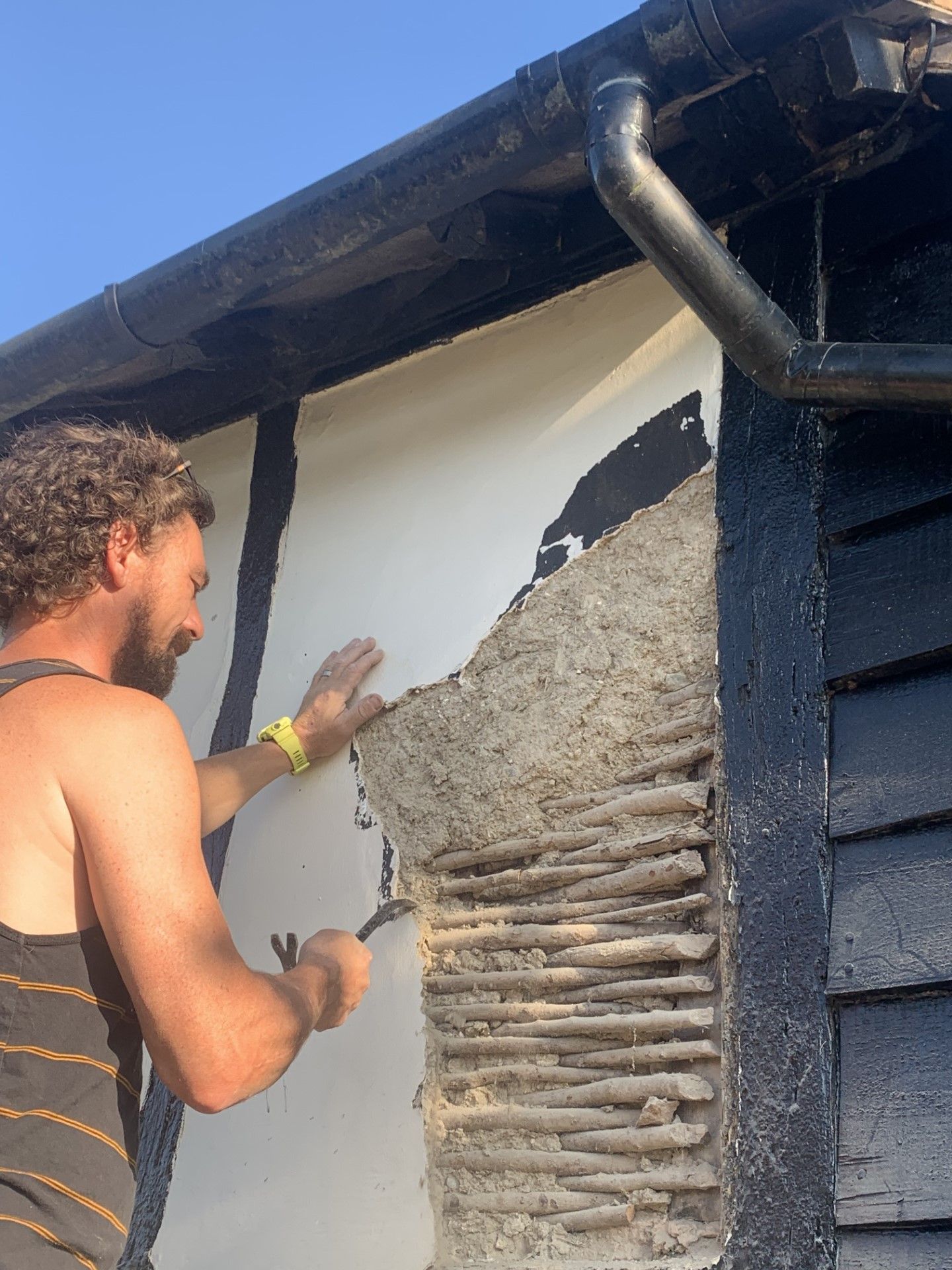 Man removing plaster from a wall with a tool outdoors. Building has a black painted trim.