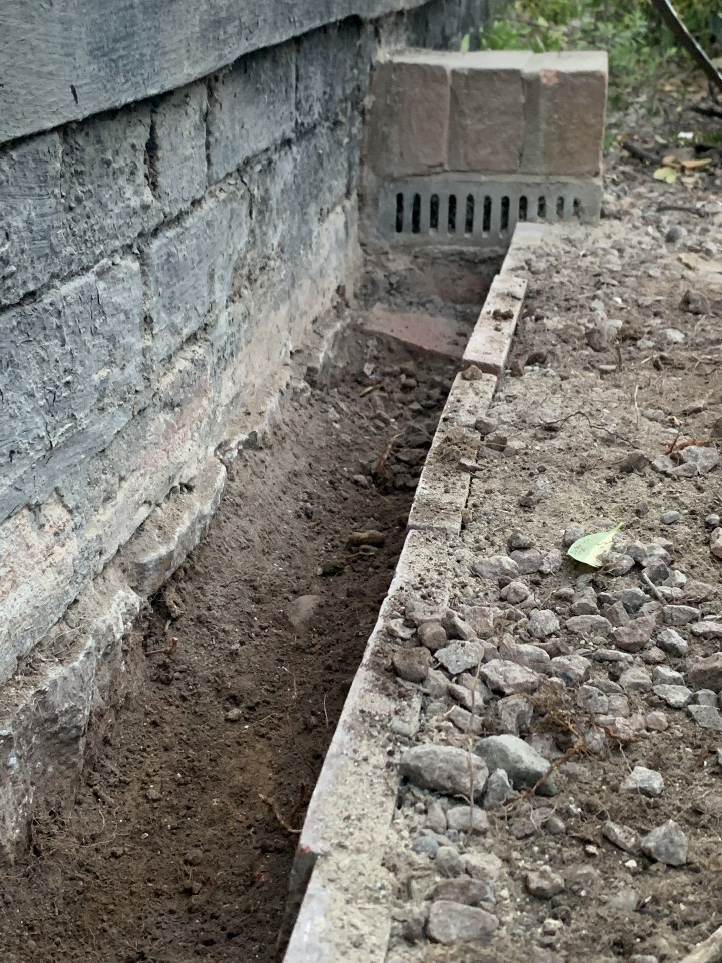 Trench dug along a brick wall, with a drainage grate at the end. Soil and debris visible.
