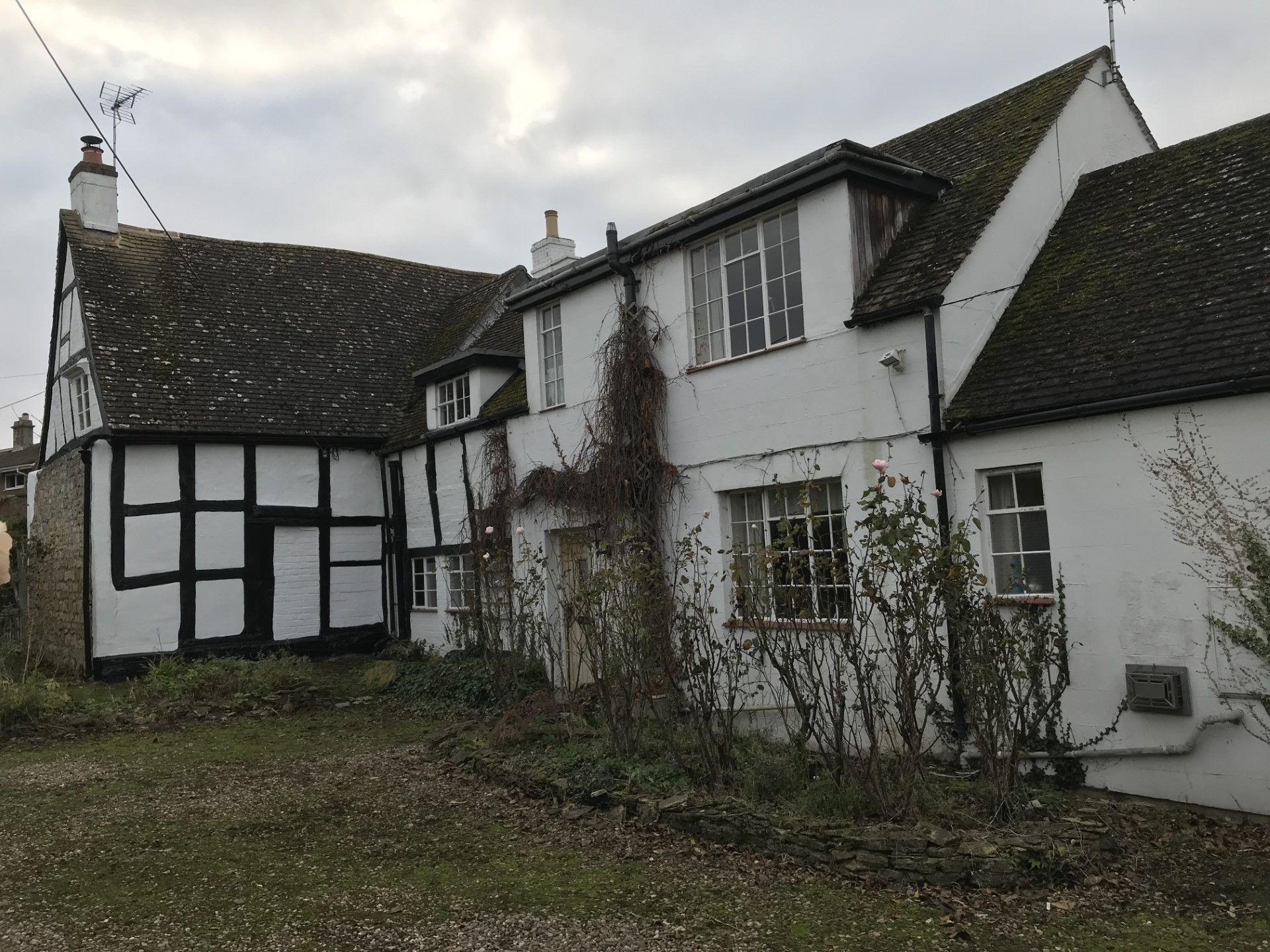 Black and white Tudor-style house with white stucco, overgrown foliage, and cloudy sky.