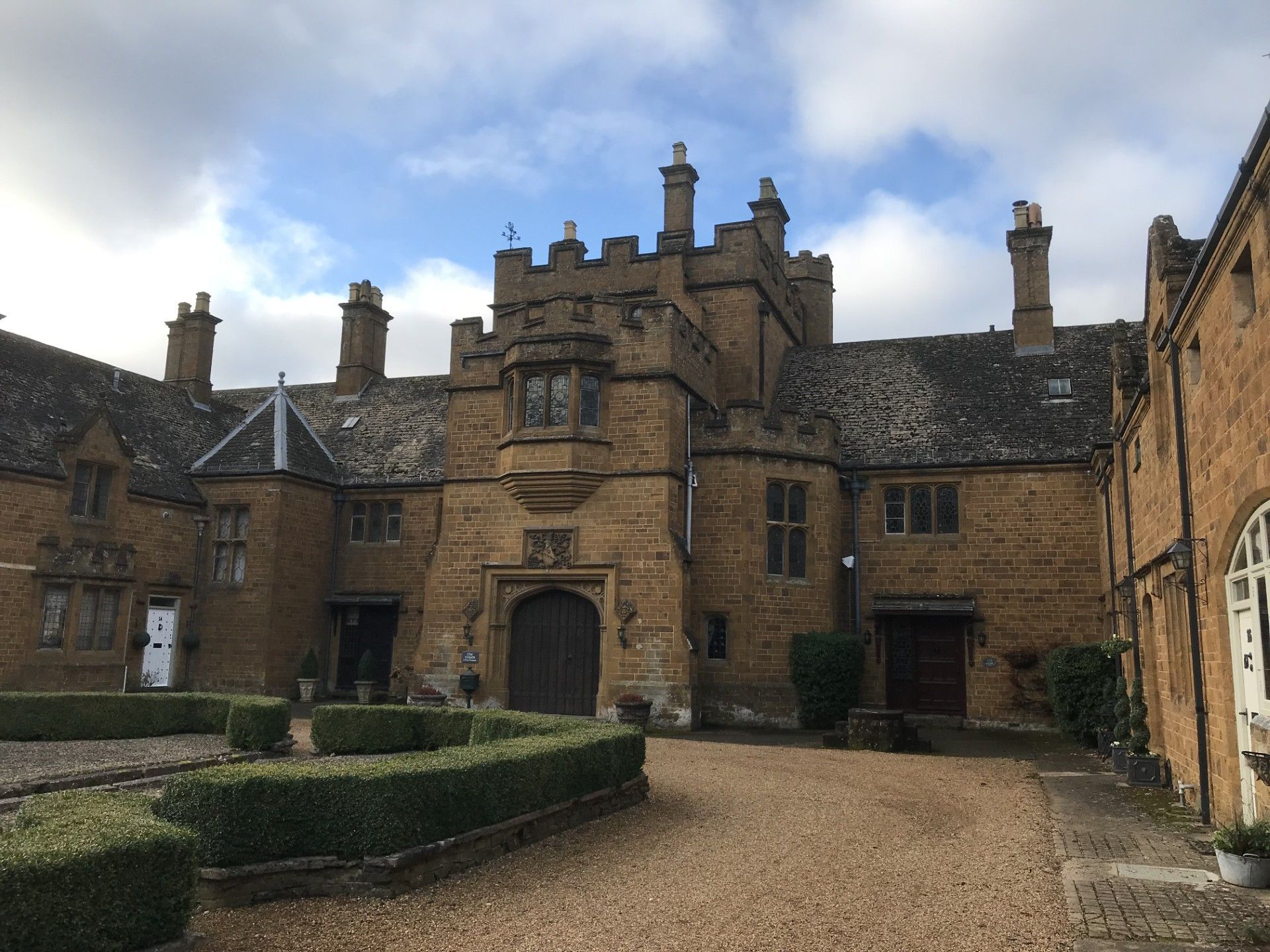 Stone manor house with arched entrance, courtyard, and low hedges against a cloudy sky.
