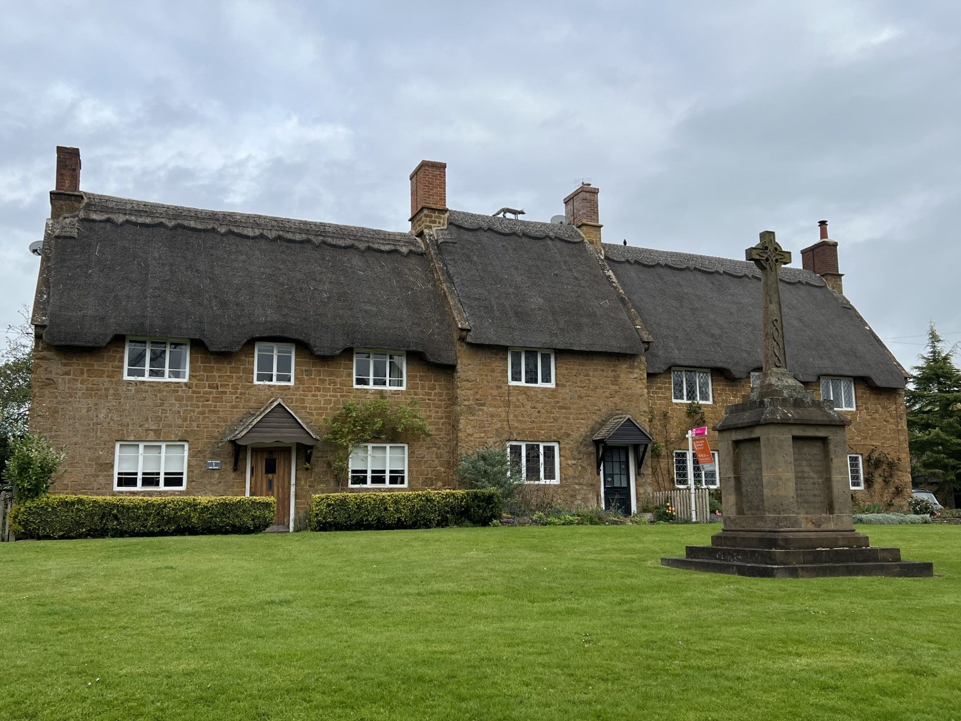Thatched-roof cottages with stone cross monument on a green lawn under cloudy sky.
