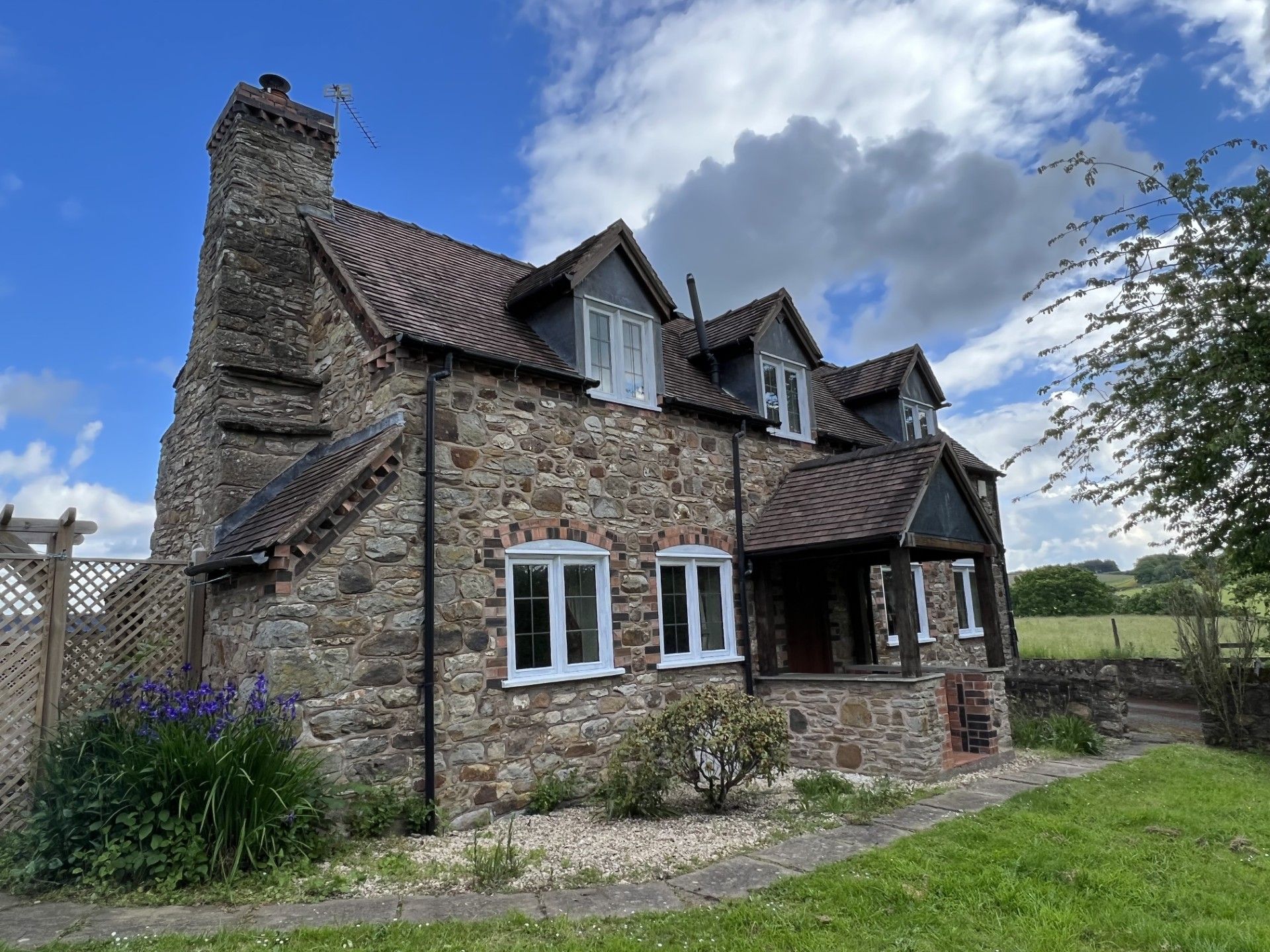 Stone cottage with chimney and dormers under a cloudy sky, with porch and garden.