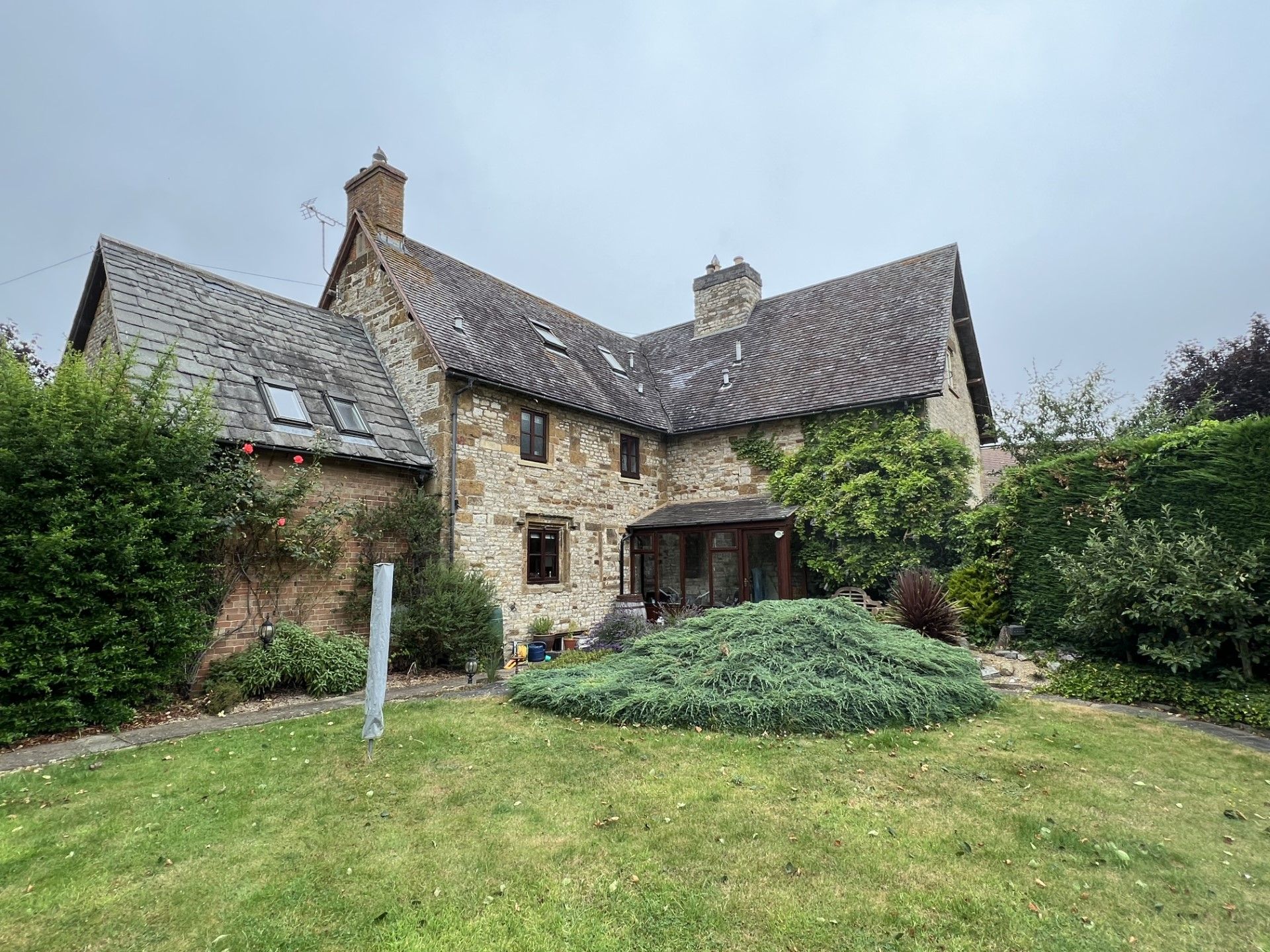 Stone cottage with a grass yard, bushes, and overcast sky.