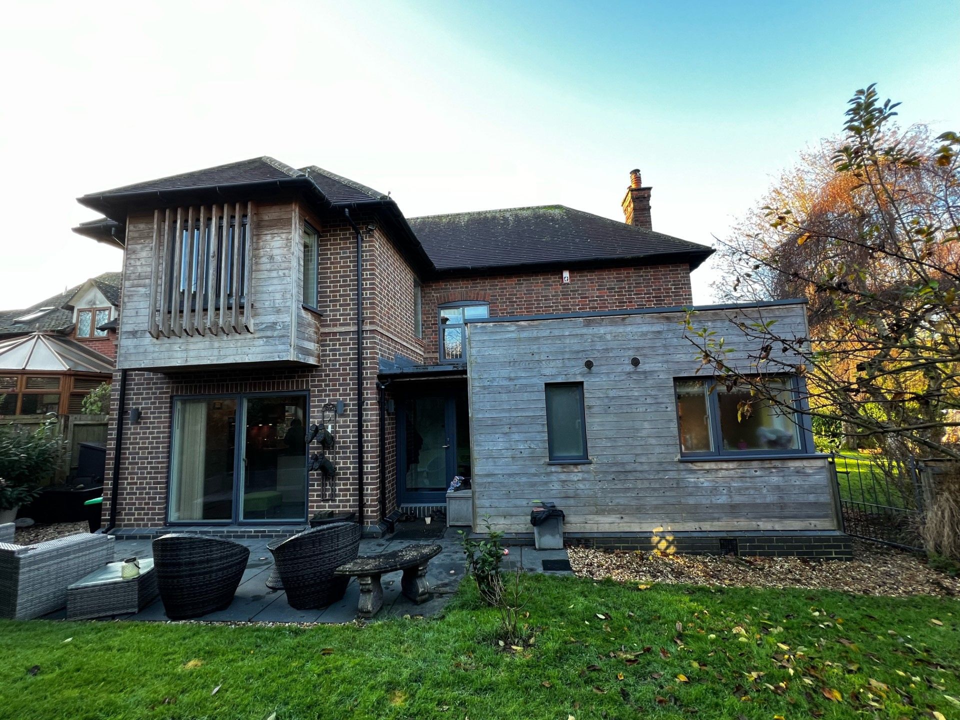 A two-story brick house with a wooden extension. Large windows and a garden visible.