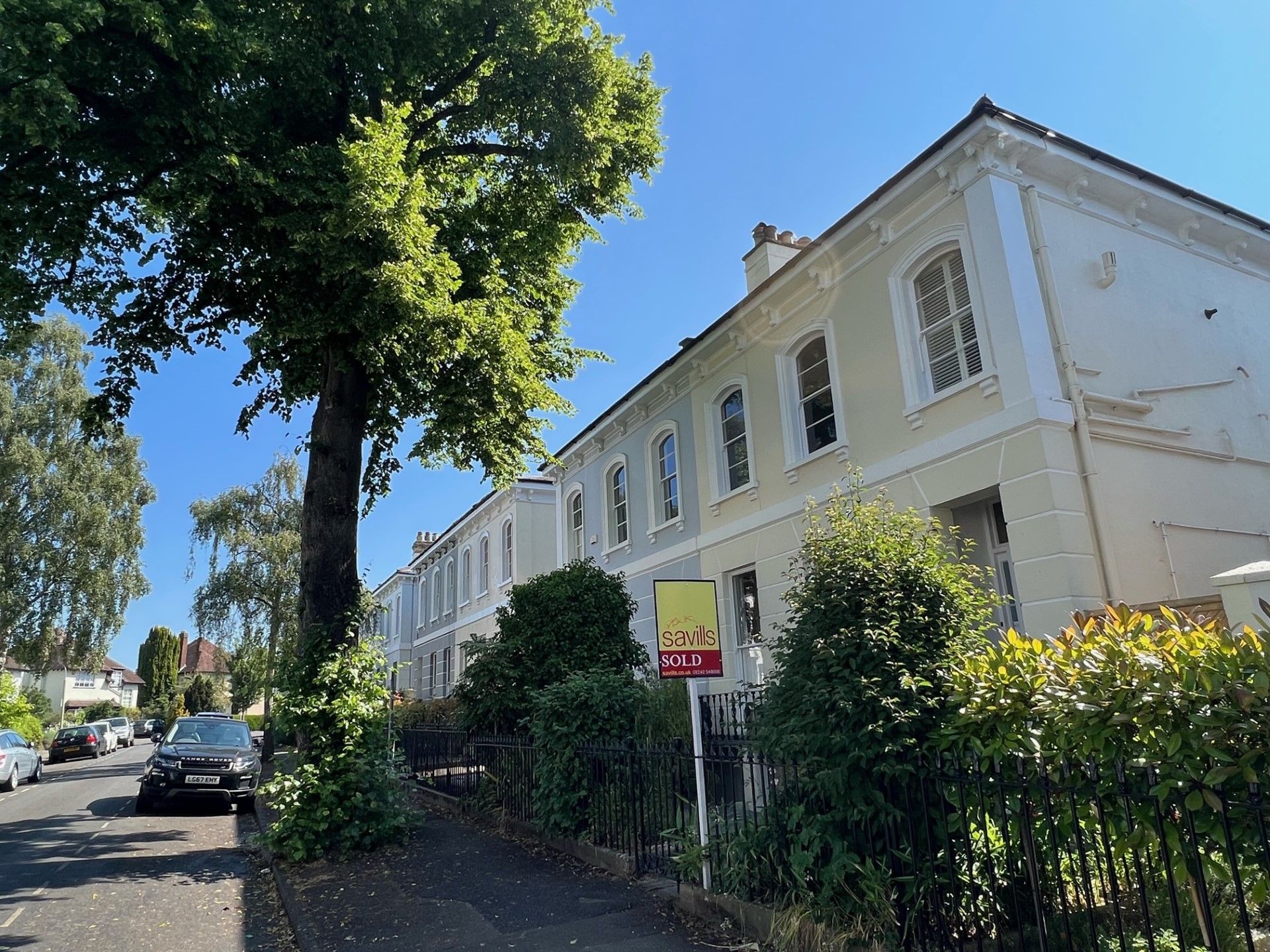 Row of light-colored townhouses with trees and a for sale sign on a sunny street.