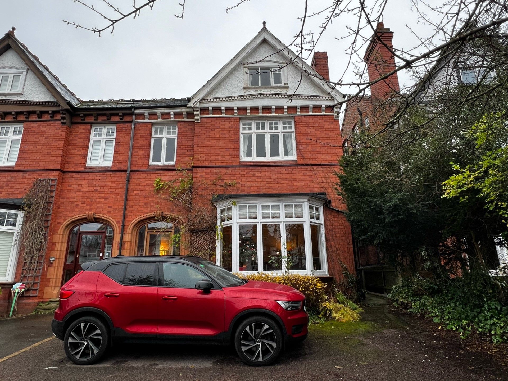Red Volvo SUV parked in front of a brick house with white trim on a cloudy day.