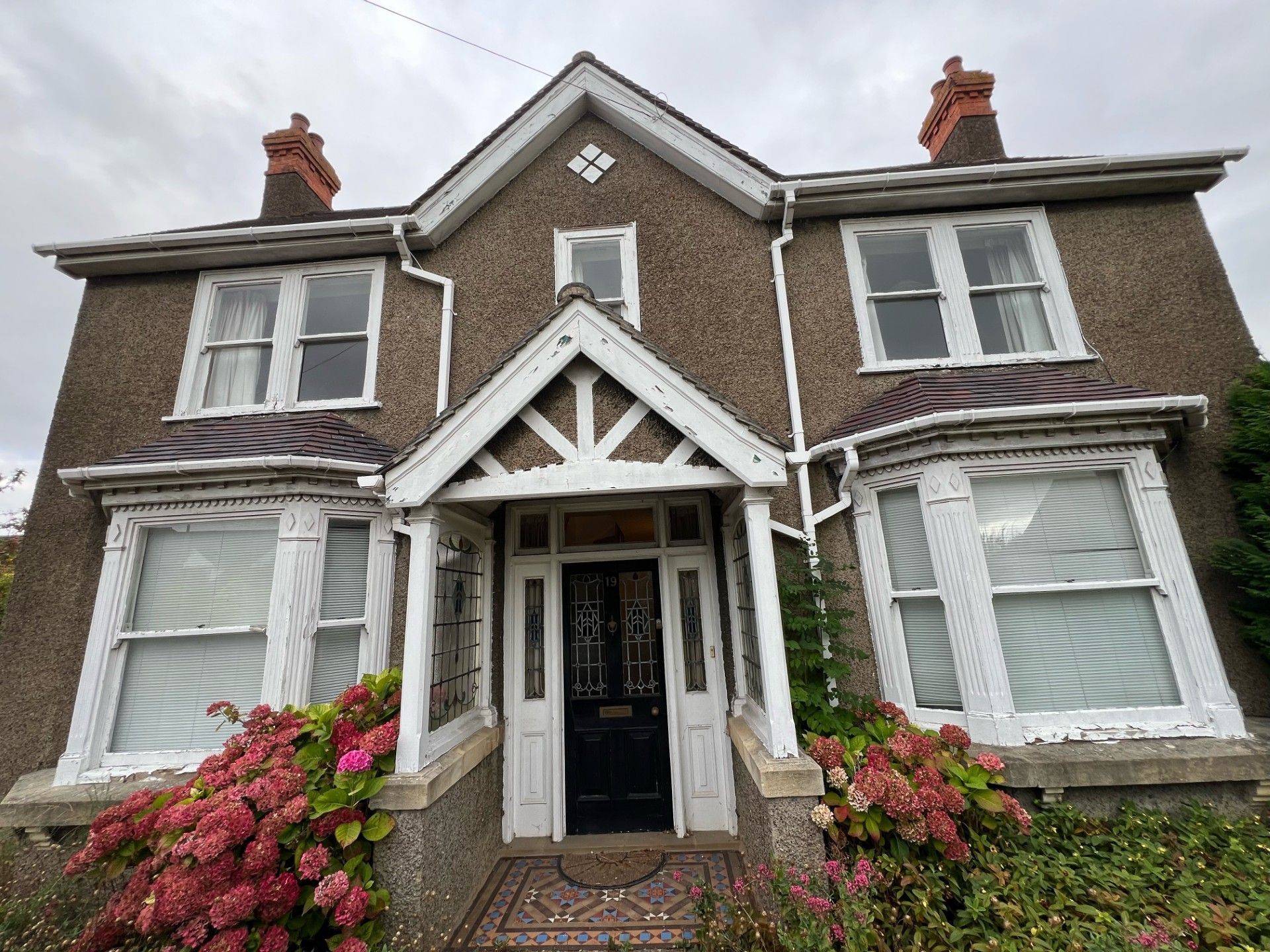 Two-story brown house with white trim, bay windows, and a porch, with red flowers.