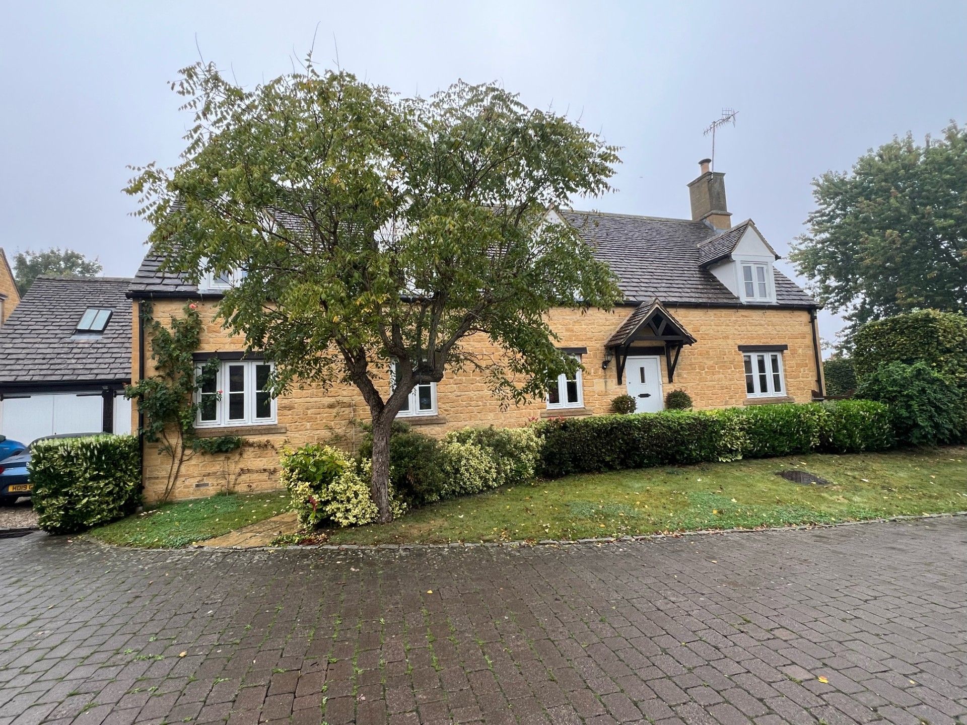 Yellow brick cottage with white windows, door, and green shrubbery. Cloudy day.