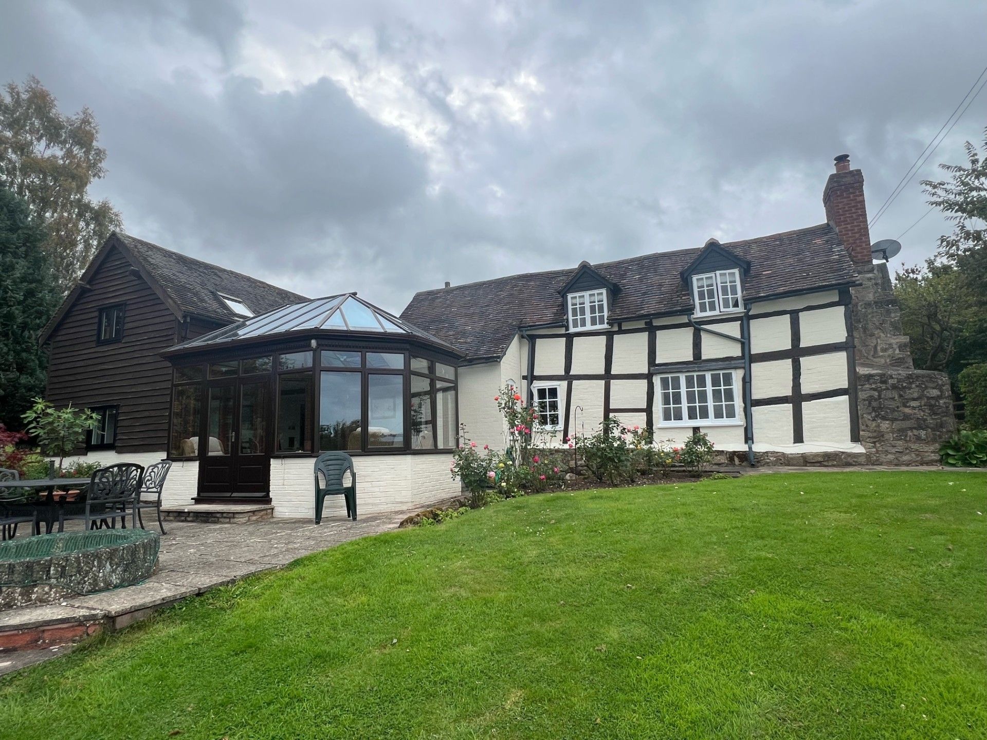 A traditional English cottage with a white and black facade, glass sunroom, and green lawn under a cloudy sky.