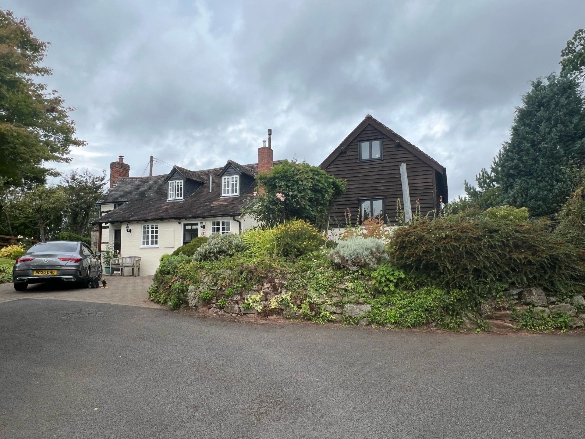 Cottage with stone facade, dark wooden extension, and car parked on the drive under an overcast sky.