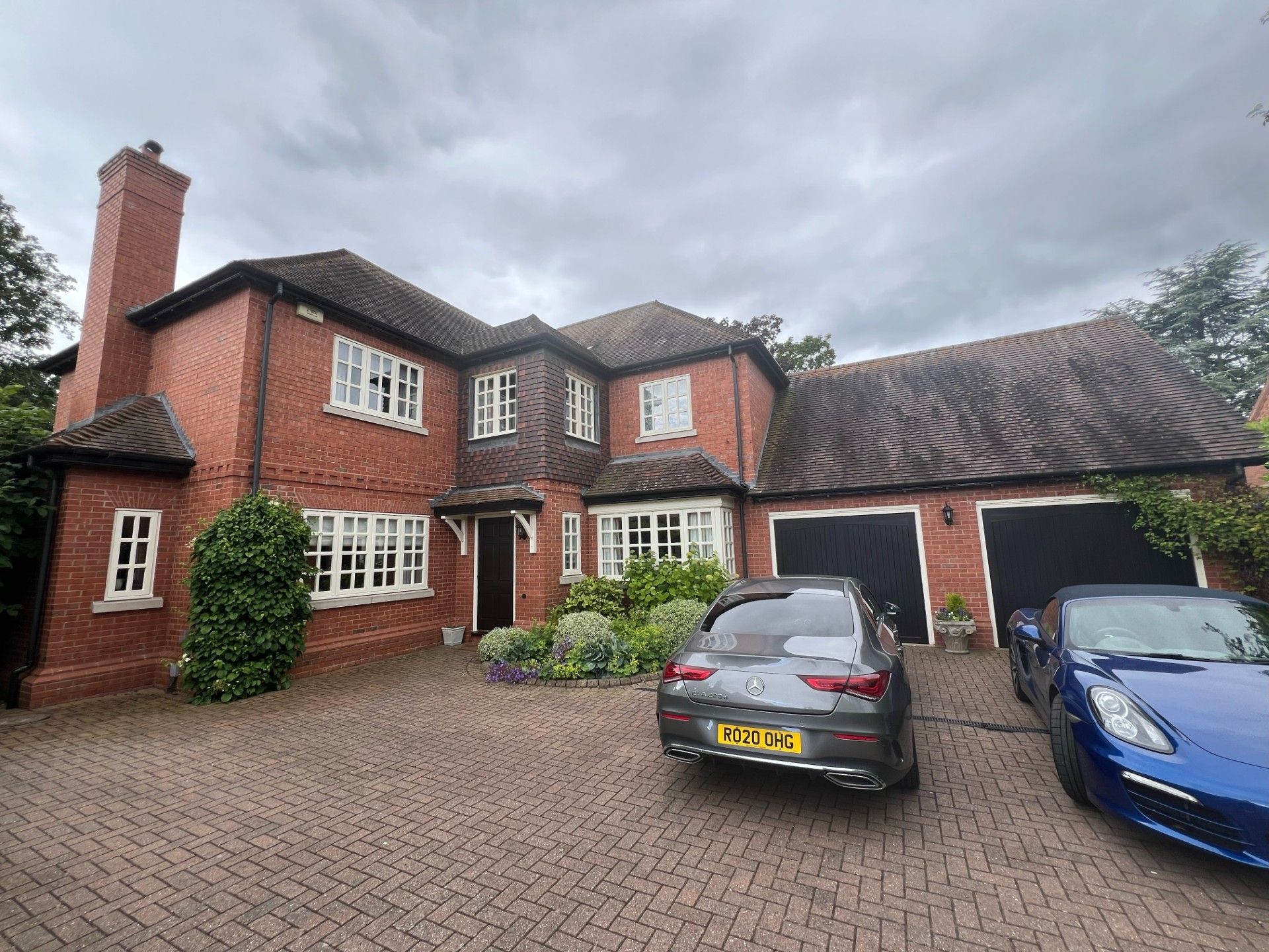Red brick house with a driveway and two cars parked in front. Overcast sky.