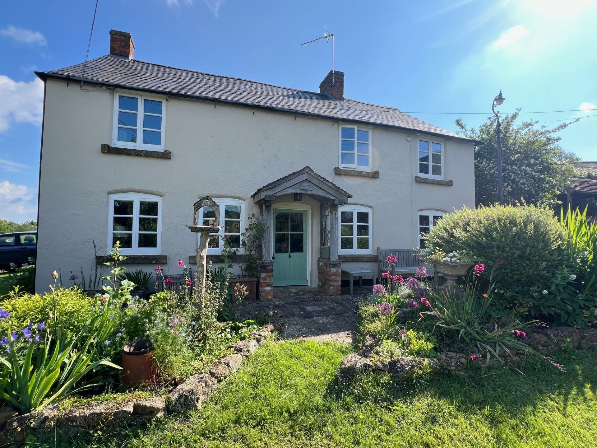Cottage with a green door and white-framed windows, surrounded by a garden on a sunny day.