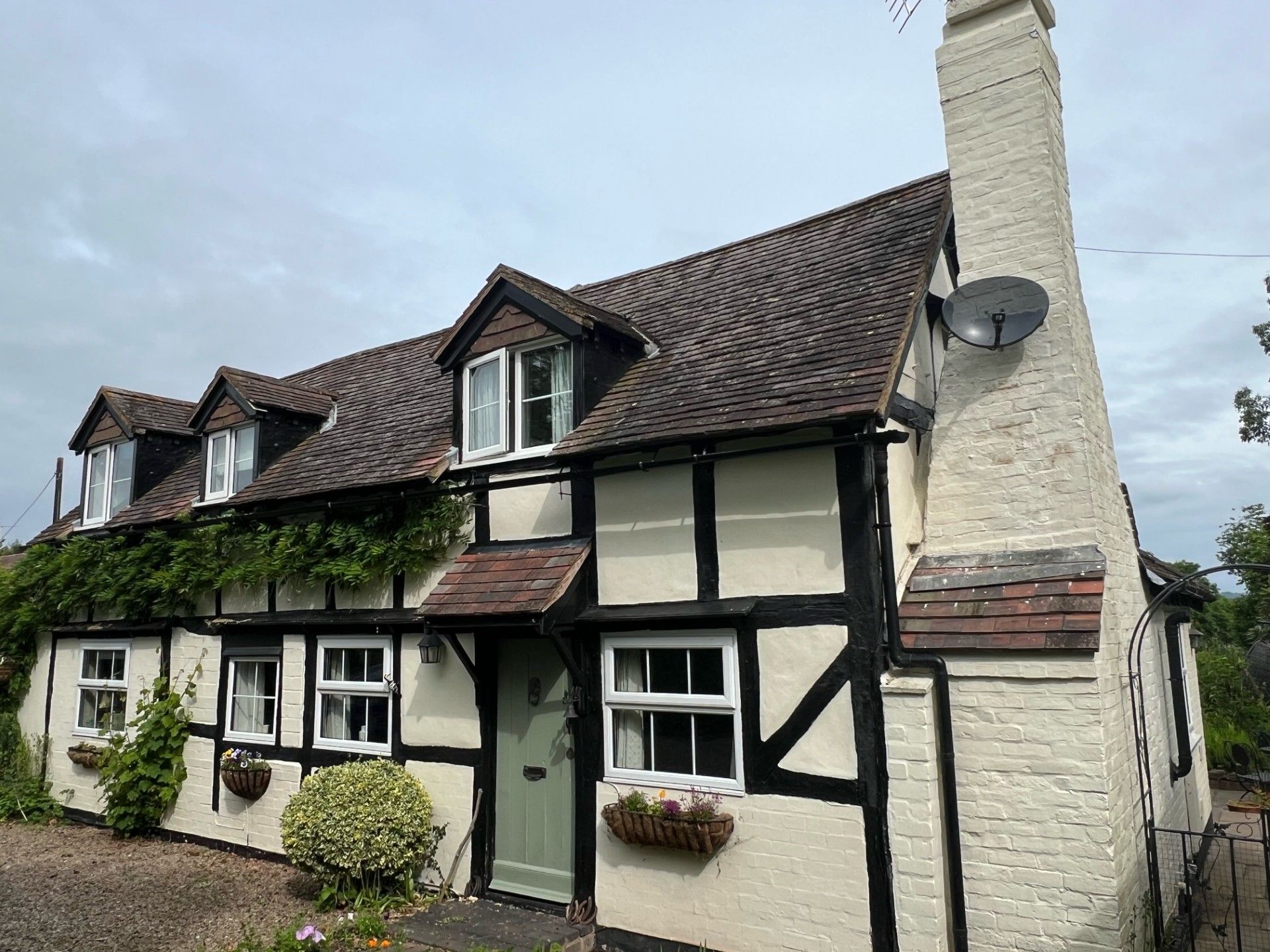 Cottage with black timber framing, white stucco walls, and a tiled roof. Green door, dormer windows, and satellite dish.