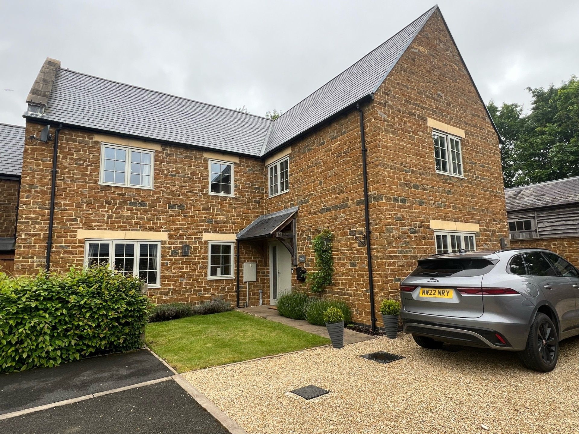 Two-story house with yellow brick exterior, gray roof, gravel driveway, and a silver car parked outside.