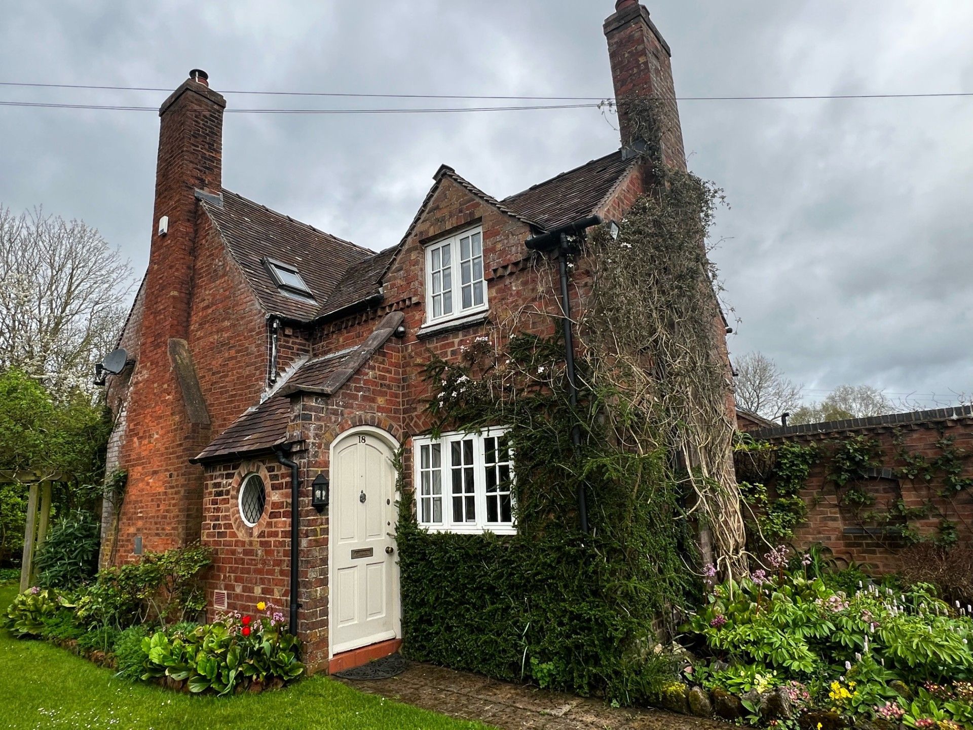 Brick cottage with white door, windows, and climbing greenery; cloudy sky.